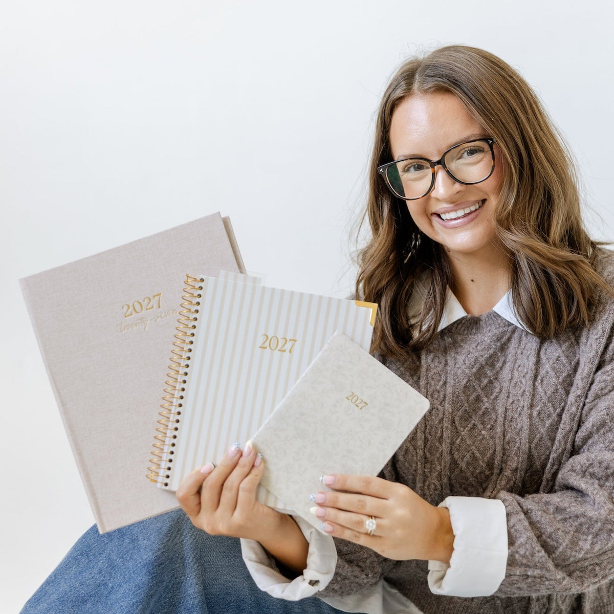 A smiling young woman with glasses sits on a white floor holding three spiral-bound 2025 planners. She is wearing a brown cable-knit sweater over a white collared shirt and blue jeans against a clean white background.