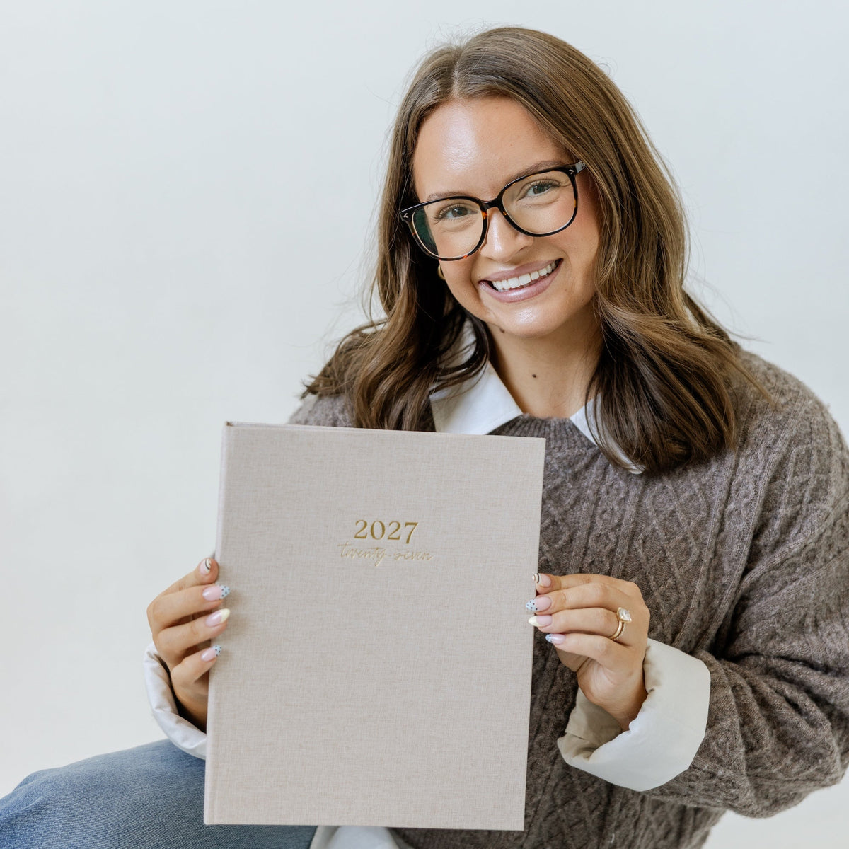 A smiling woman with glasses and brown hair sits against a light gray background, holding up a beige 2027 planner or calendar with gold text.