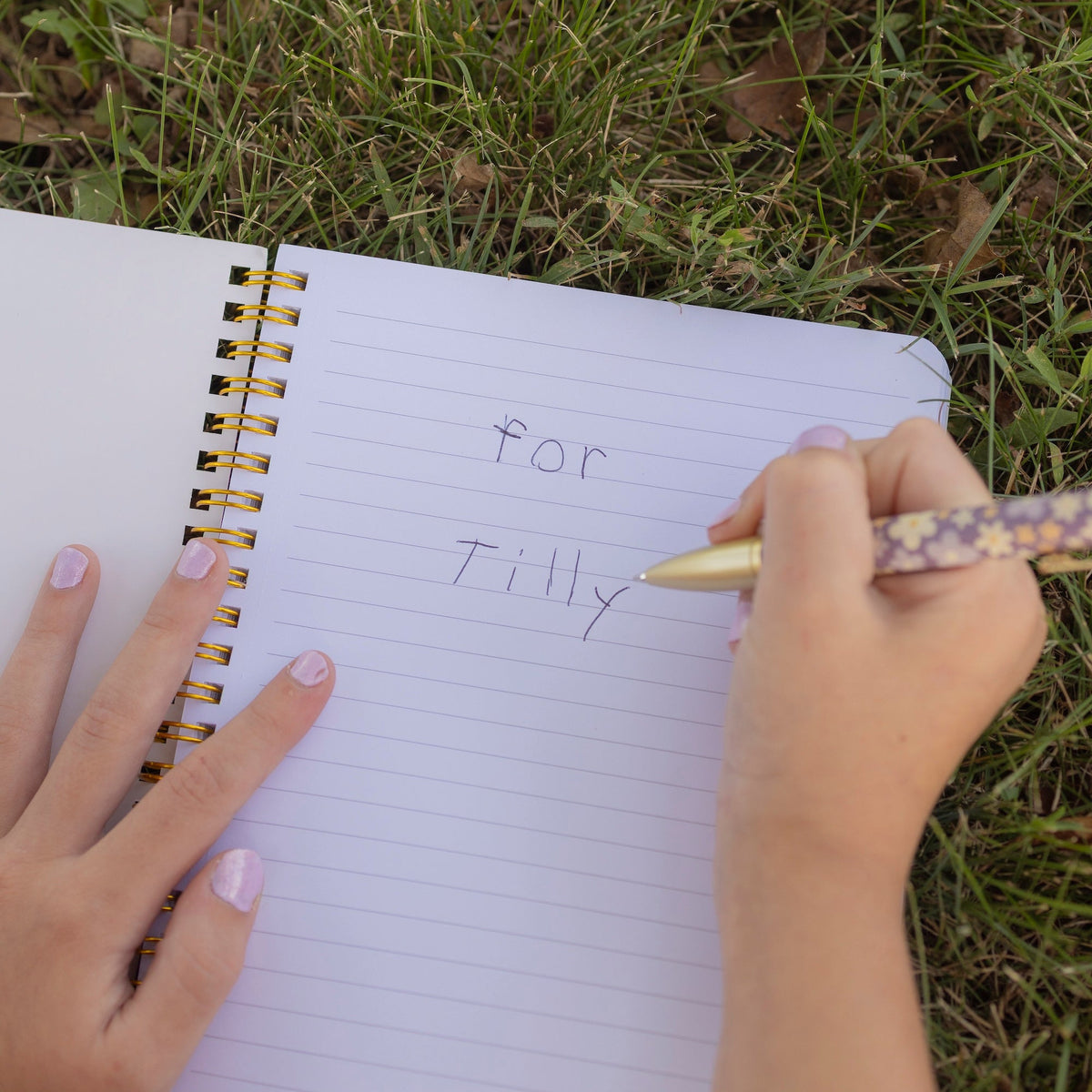 A close-up of a person writing in a lined spiral notebook on grass, with the handwritten text 'for Tilly' visible and a patterned pen held in the right hand.