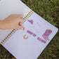 A hand is placing a small sticker on a lined notebook lying on grass. The notebook has western-themed stickers including a purple hat, purple boots, a gold horseshoe, and the words 'For Tilly Groom Up Yeehaw!' written in purple text.