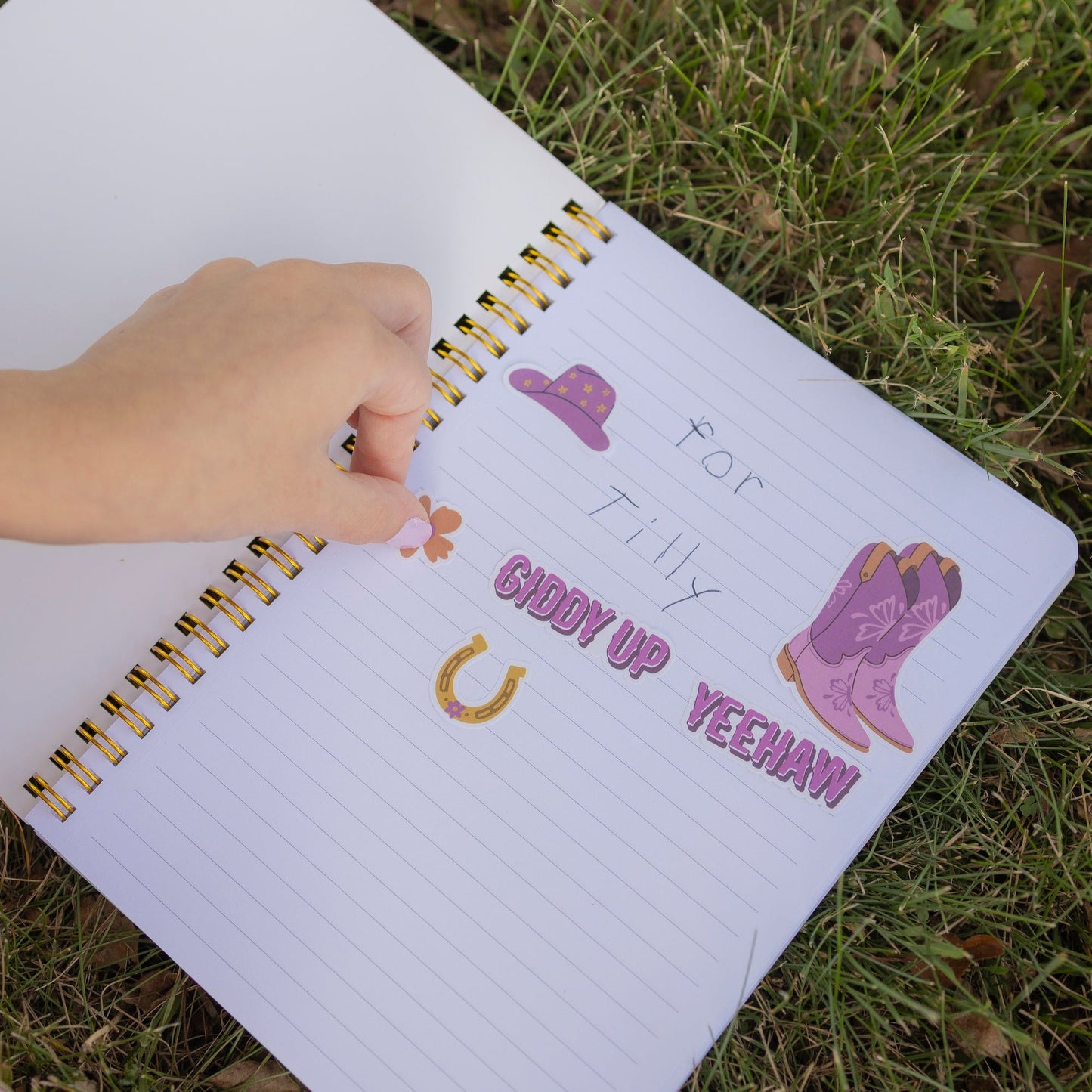 A hand is placing a small sticker on a lined notebook lying on grass. The notebook has western-themed stickers including a purple hat, purple boots, a gold horseshoe, and the words 'For Tilly Groom Up Yeehaw!' written in purple text.