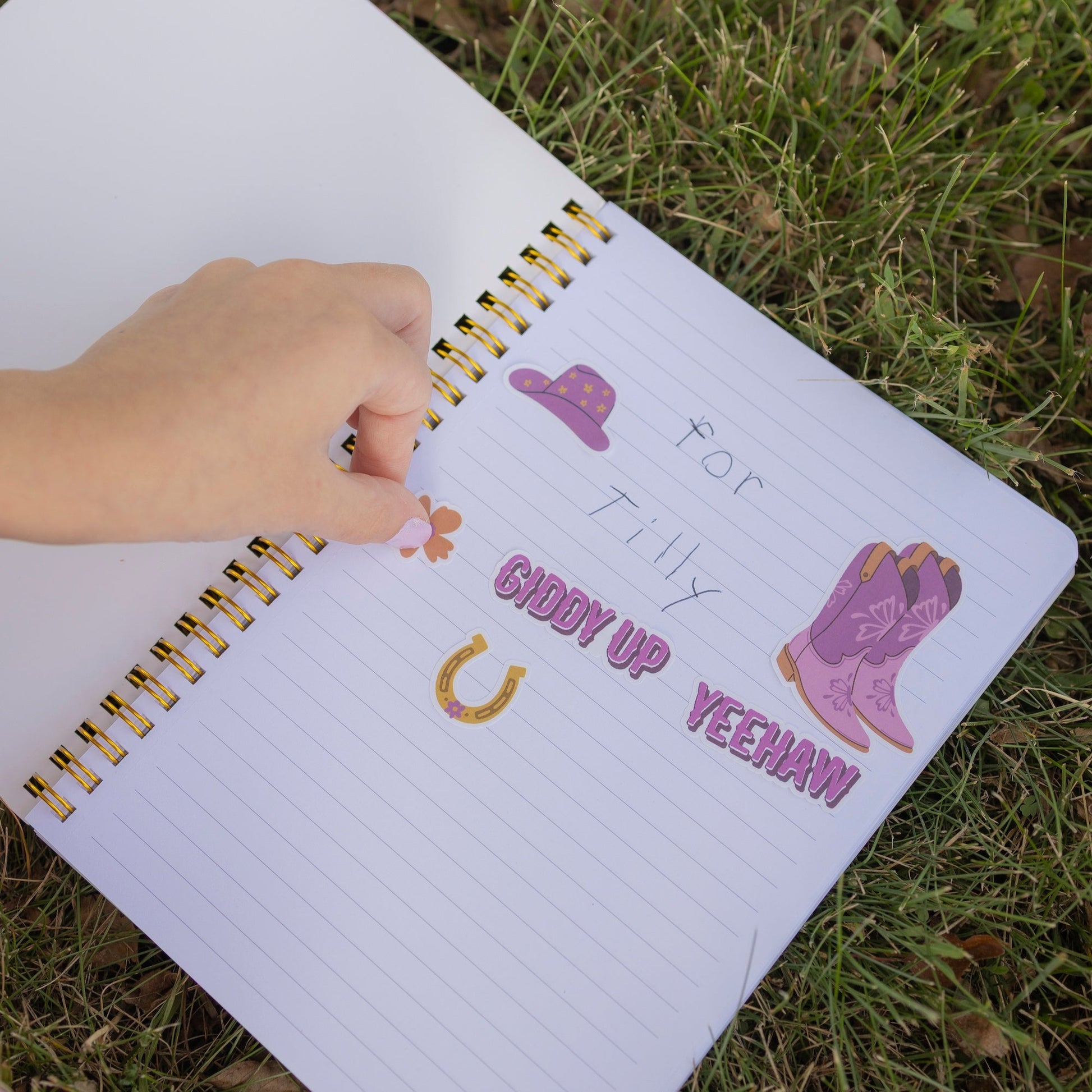 A hand is placing a small sticker on a lined notebook lying on grass. The notebook has western-themed stickers including a purple hat, purple boots, a gold horseshoe, and the words 'For Tilly Groom Up Yeehaw!' written in purple text.