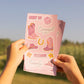 A person outdoors holds up a stack of pink cowboy-themed sticker sheets. The stickers feature pink cowboy boots, floral designs, horseshoes, and phrases like 'Crazy Horse' and 'Yeehaw Heydy' on a soft pink background.