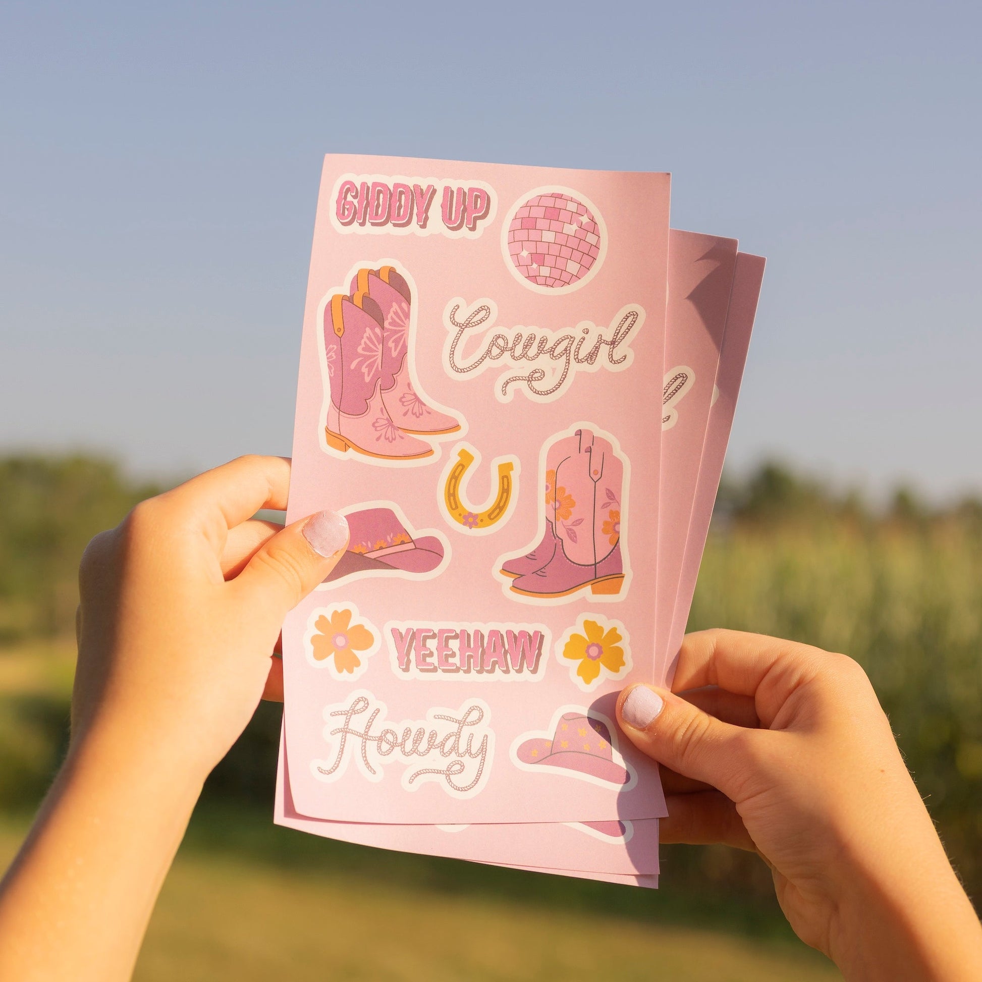 A person outdoors holds up a stack of pink cowboy-themed sticker sheets. The stickers feature pink cowboy boots, floral designs, horseshoes, and phrases like 'Crazy Horse' and 'Yeehaw Heydy' on a soft pink background.