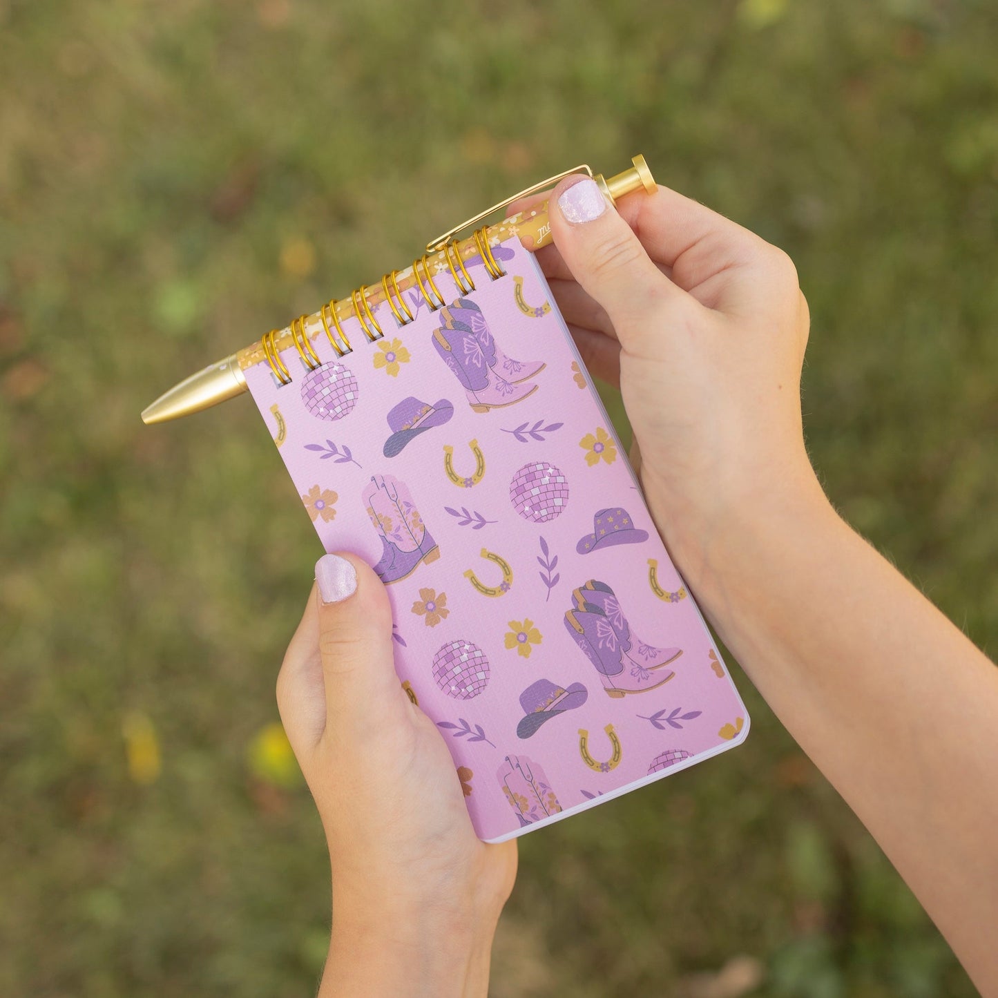 A person holding a small spiral notebook with a purple cowboy-themed pattern, including boots, hats, horseshoes, and a cowboy figure, with a gold pen in one hand against a blurred grassy background.