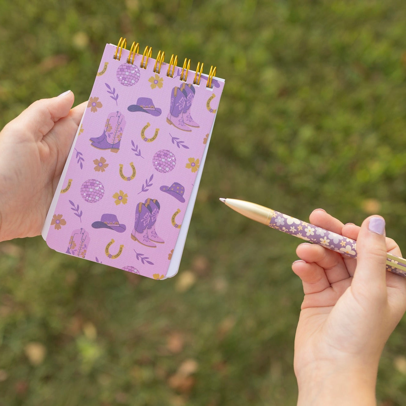 A person holds a small spiral-bound notebook with a lavender cover decorated with purple cowboy boots, gold horseshoes, and other western-themed illustrations, while holding a matching patterned pencil in the other hand against a blurred green outdoor background.