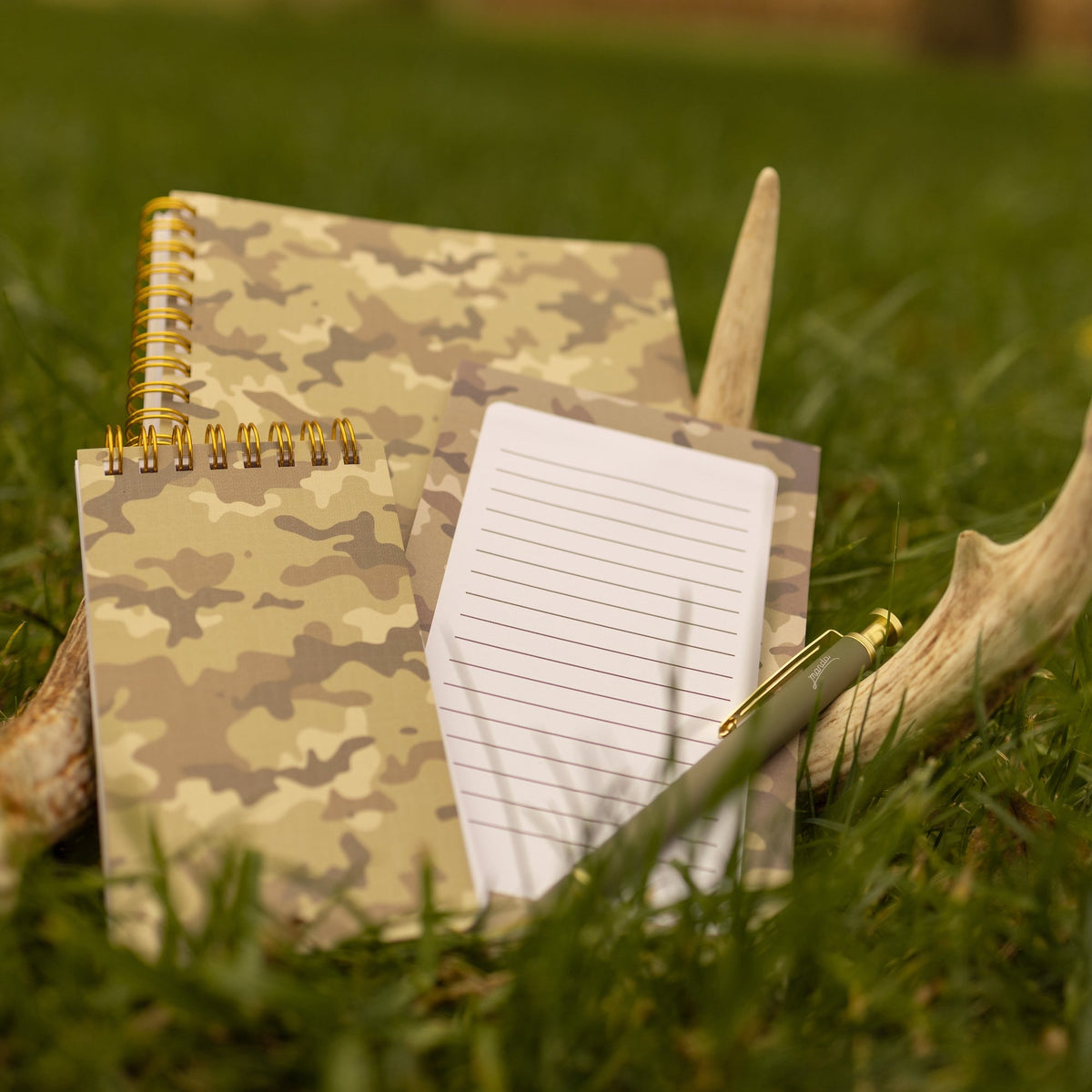 A camouflage-patterned spiral notebook and a lined notepad with a gold pen resting on a pair of antlers in a grassy outdoor field.