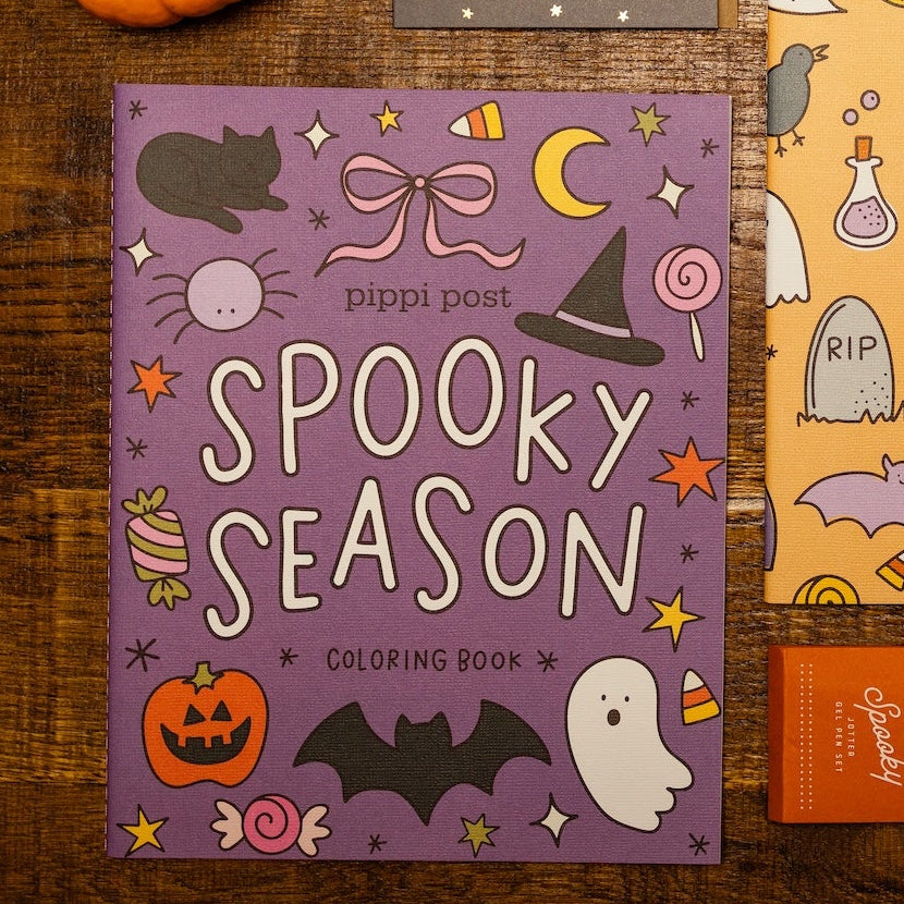 Two illustrated coloring books for Halloween displayed on a rustic wooden surface with small pumpkins, two black pens, and an orange pencil case. One book cover is purple and reads 'Spooky Season,' and the other is peach/orange and reads 'Hocus Pocus.
