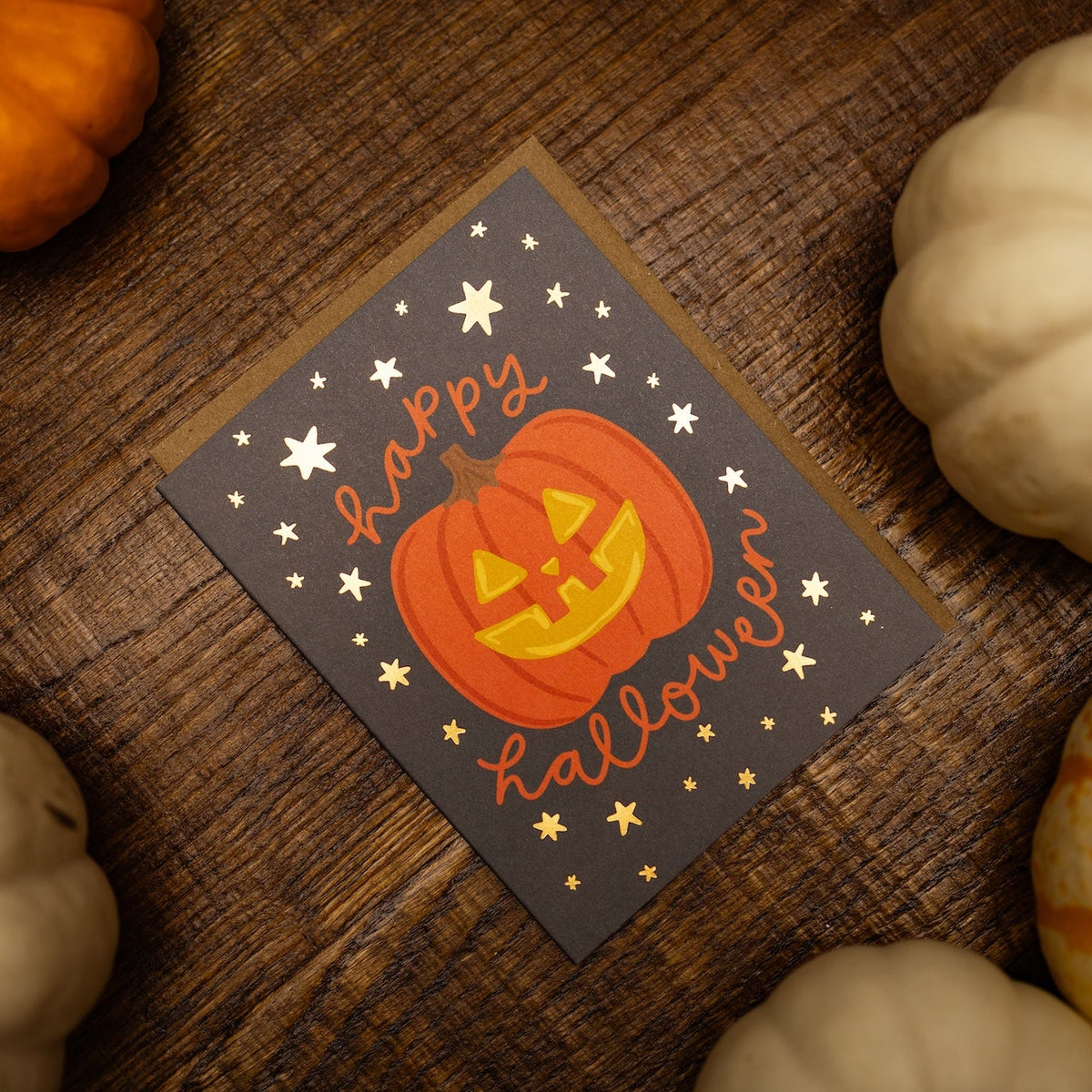 A square Halloween greeting card with a jack-o'-lantern illustration and the words 'Happy Halloween' in orange script, placed on a rustic wooden surface surrounded by small pumpkins.