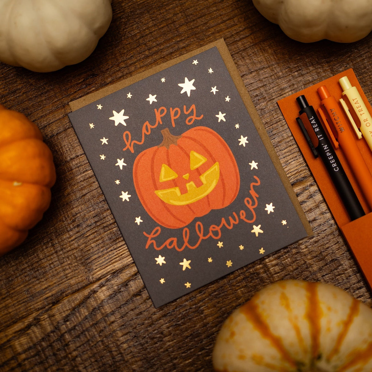 A Halloween greeting card with a jack-o'-lantern and the words 'happy Halloween' in orange script, styled on a rustic wooden surface with small pumpkins around it. Beside the card is an orange stationery gift set containing pens and a pencil in an orange box labeled 'The Spooky Stationery Gift Set'.