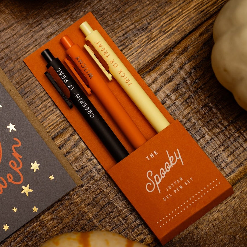 A styled flat-lay of a 'Happy Halloween' greeting card next to a matching orange pen box labeled 'The Spooky'. The scene is decorated with small pumpkins on a rustic wooden surface.