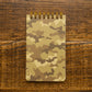A spiral-bound notepad with a tan and brown camouflage pattern, placed on a dark wood tabletop.