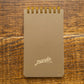 A vertical spiral-bound notebook with a textured taupe cover and gold wire binding, featuring the brand name 'manda' in white script near the bottom, set against a rustic wooden background.