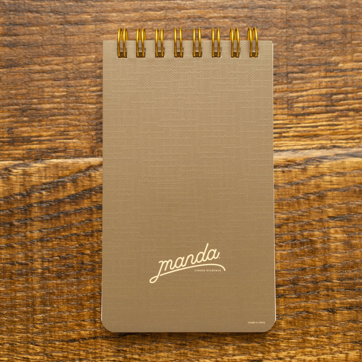 A vertical spiral-bound notebook with a textured taupe cover and gold wire binding, featuring the brand name 'manda' in white script near the bottom, set against a rustic wooden background.