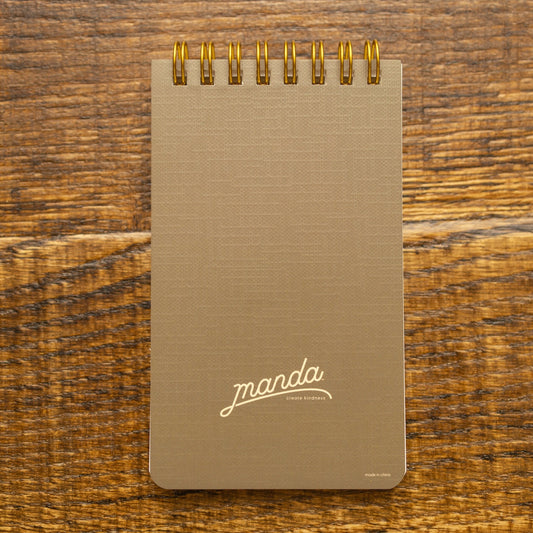 A vertical spiral-bound notebook with a textured taupe cover and gold wire binding, featuring the brand name 'manda' in white script near the bottom, set against a rustic wooden background.
