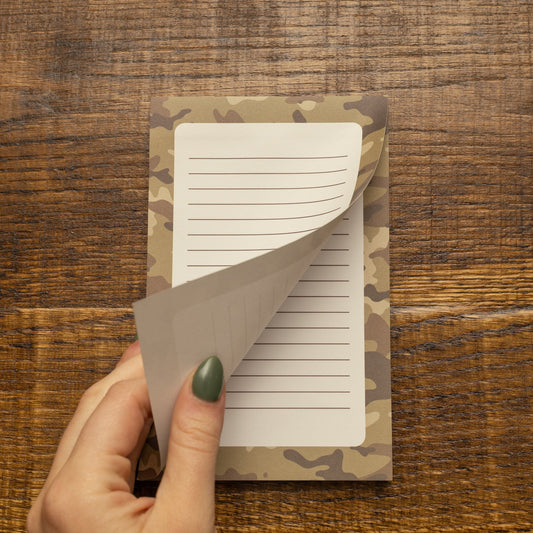 A hand with green nail polish peels back a lined paper page from a notepad featuring a camouflage pattern, set against a dark wood background.