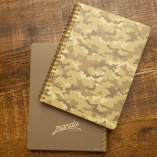 Two spiral-bound notebooks on a rustic wooden table. The top notebook features a beige and brown camouflage pattern, while the bottom notebook is solid taupe with the brand name 'marcha' printed in white script.