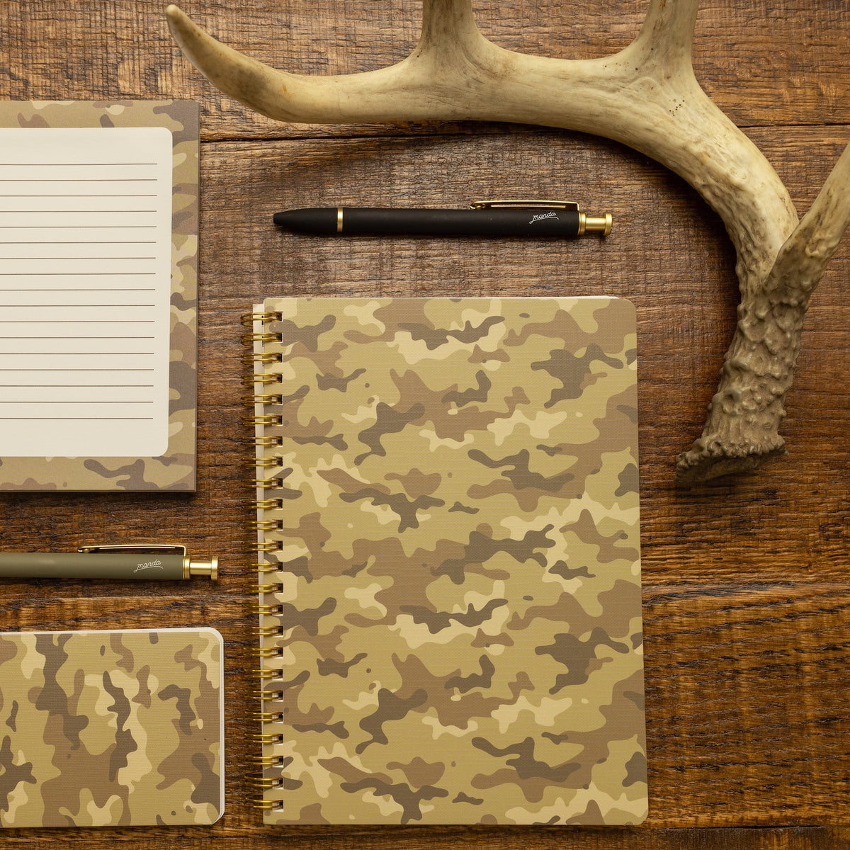 A coordinated set of camouflage stationery items arranged on a wooden tabletop, including a lined notepad, a spiral notebook, a small notebook, two pens, and a deer antler prop.