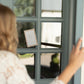 A person in a white lace top stands in front of a painted blue-gray French door with multiple glass panes, holding a small card against the glass.