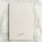 A cream-colored rectangular card or notebook cover with the name 'manda' printed in a delicate gold cursive font, placed on a textured white lace fabric background.