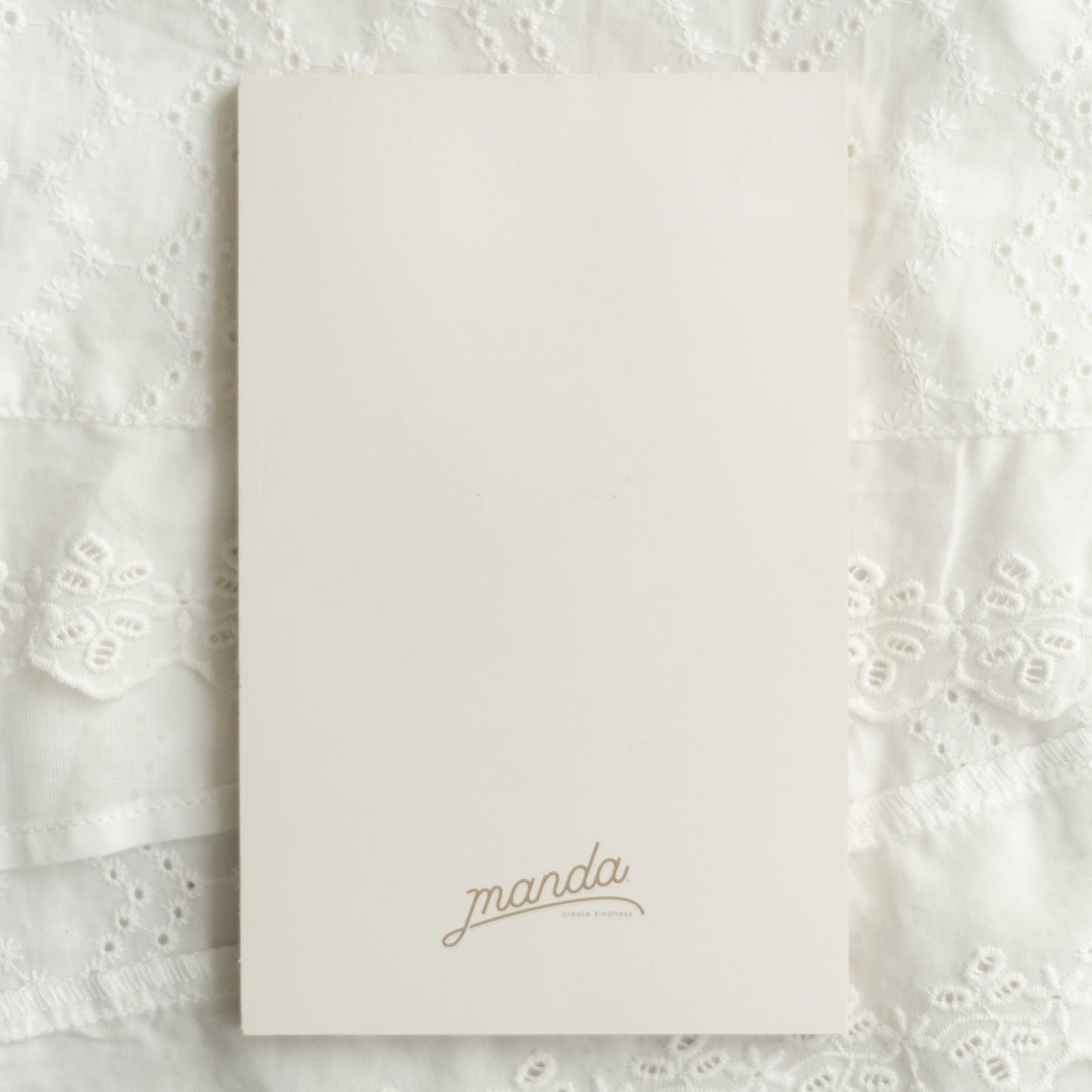 A cream-colored rectangular card or notebook cover with the name 'manda' printed in a delicate gold cursive font, placed on a textured white lace fabric background.