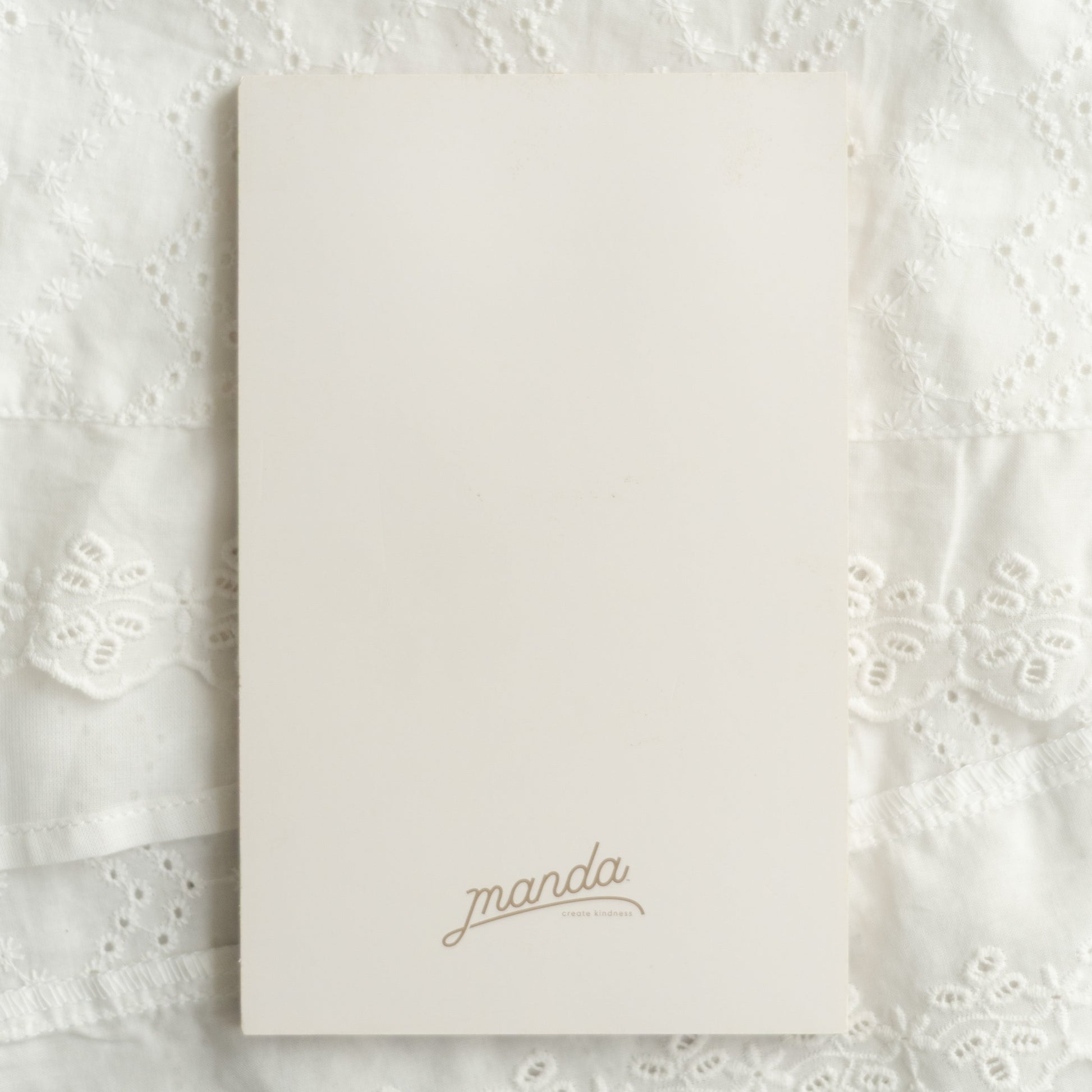 A cream-colored rectangular card or notebook cover with the name 'manda' printed in a delicate gold cursive font, placed on a textured white lace fabric background.