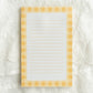 A rectangular lined notepad with a yellow and white gingham border, placed on a white embroidered lace fabric background.