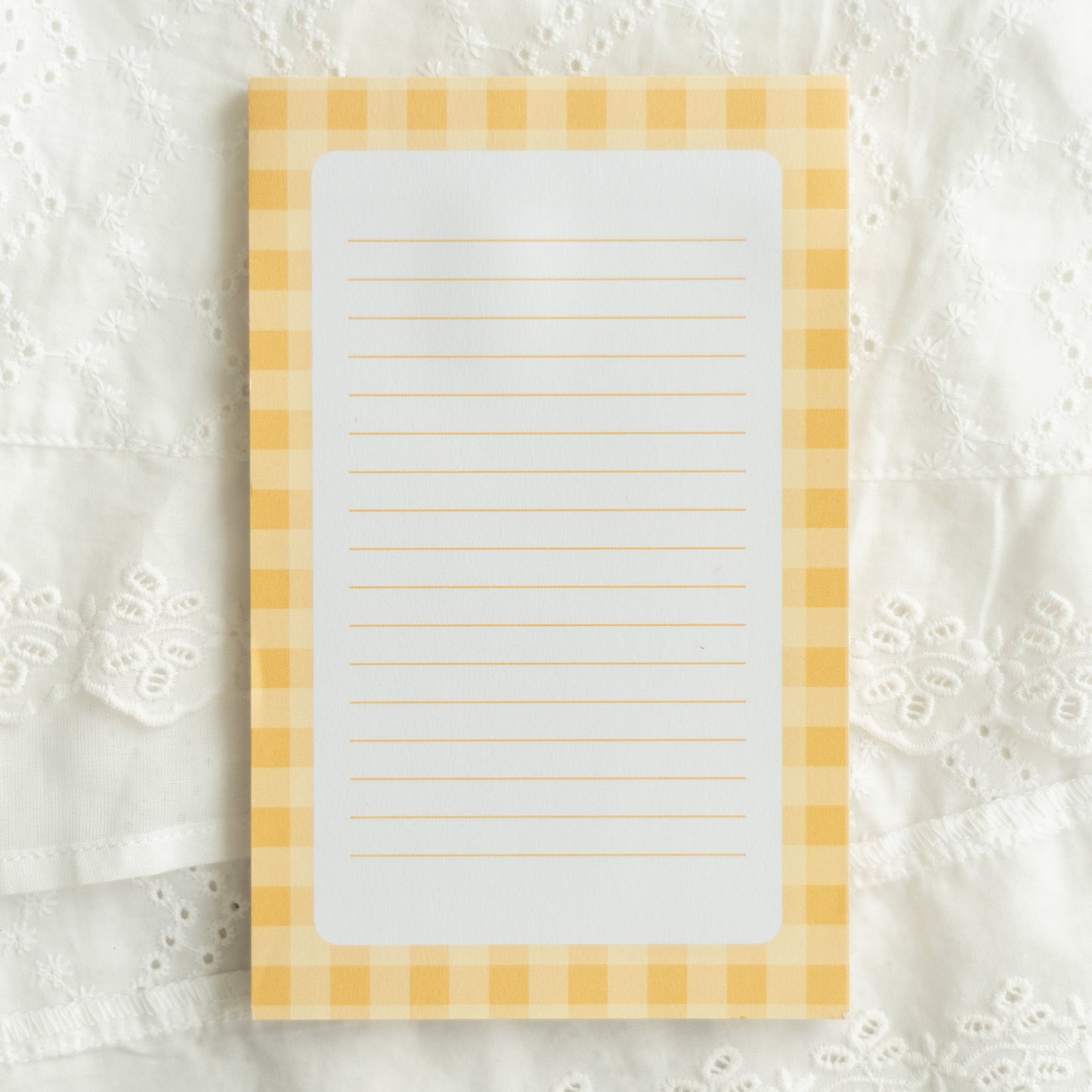 A rectangular lined notepad with a yellow and white gingham border, placed on a white embroidered lace fabric background.