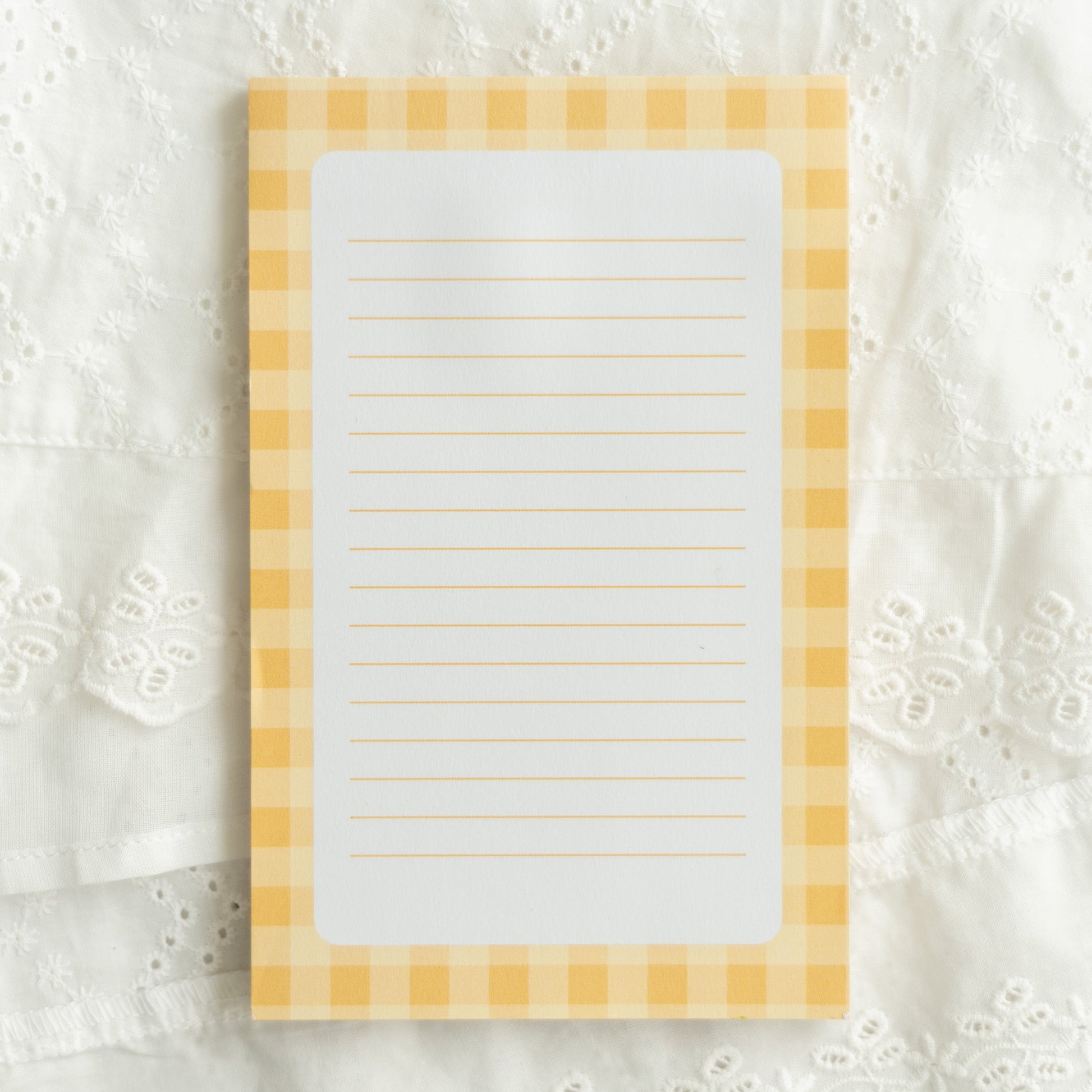 A rectangular lined notepad with a yellow and white gingham border, placed on a white embroidered lace fabric background.