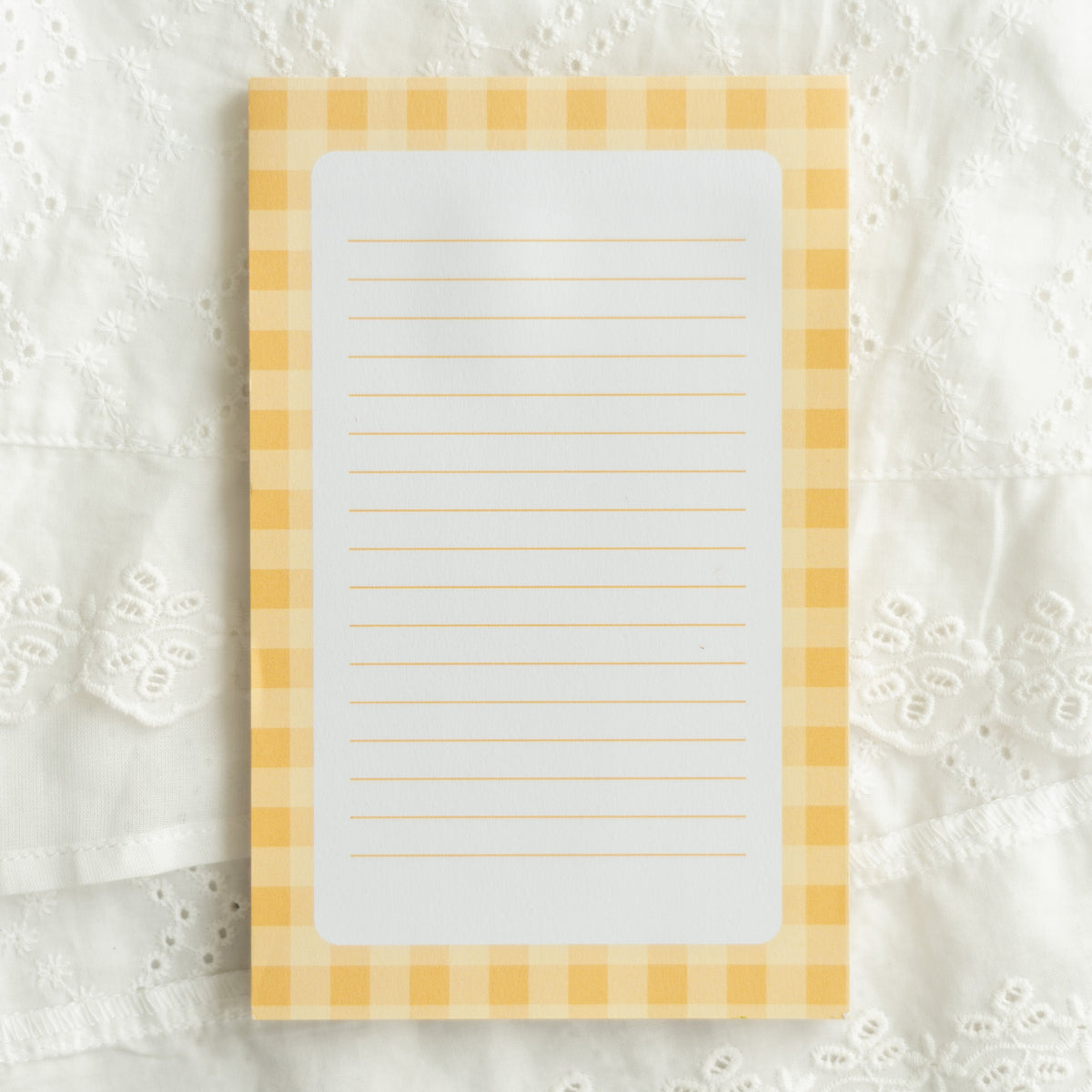 A rectangular lined notepad with a yellow and white gingham border, placed on a white embroidered lace fabric background.