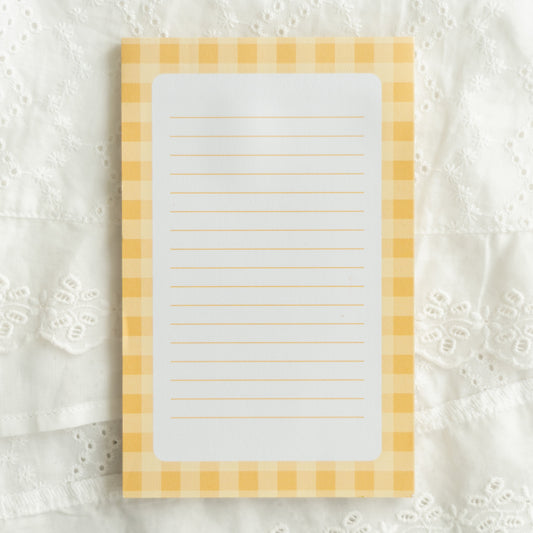 A rectangular lined notepad with a yellow and white gingham border, placed on a white embroidered lace fabric background.