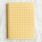 A spiral-bound notebook with a yellow and white gingham checkered cover, placed on a textured white fabric background.