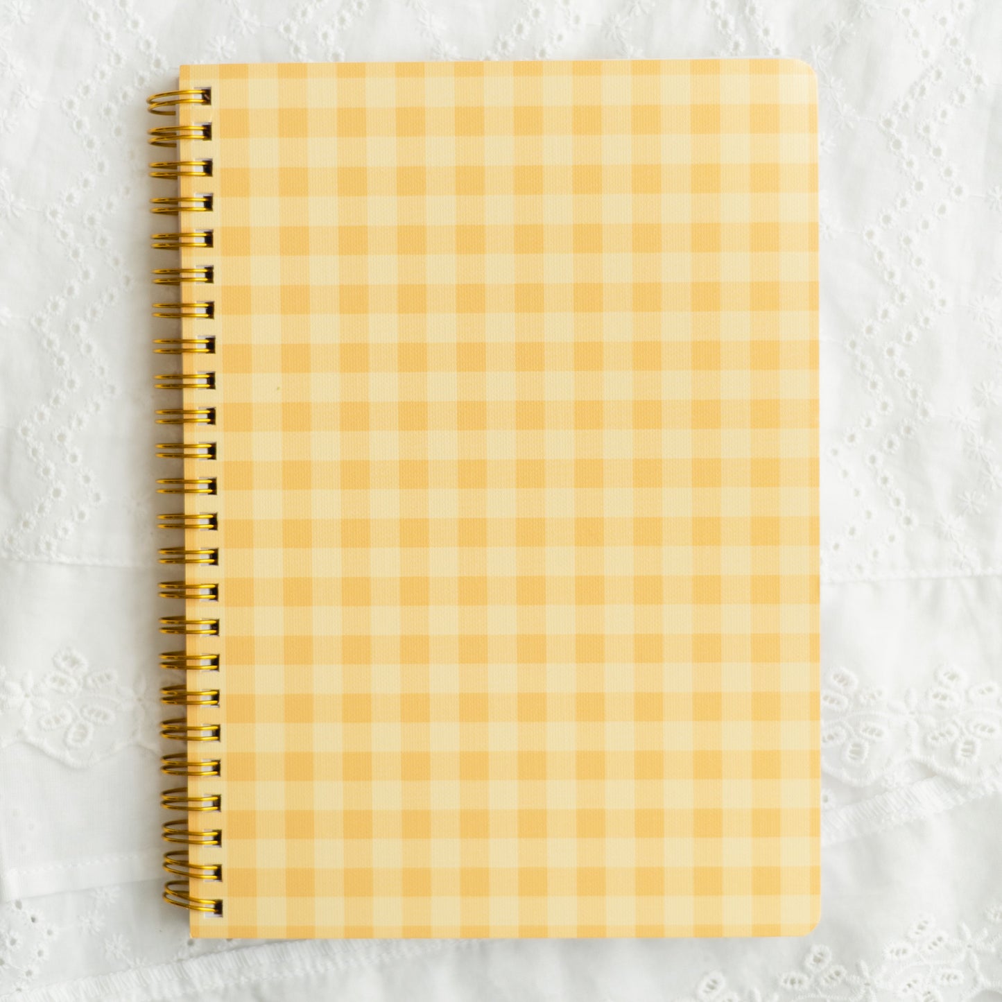A spiral-bound notebook with a yellow and white gingham checkered cover, placed on a textured white fabric background.