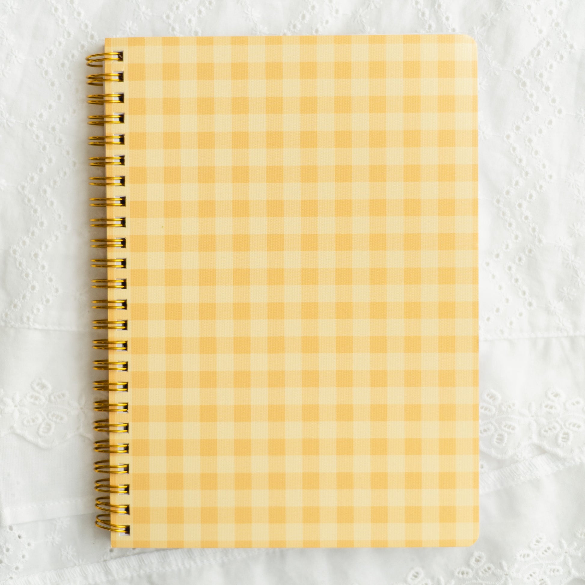 A spiral-bound notebook with a yellow and white gingham checkered cover, placed on a textured white fabric background.