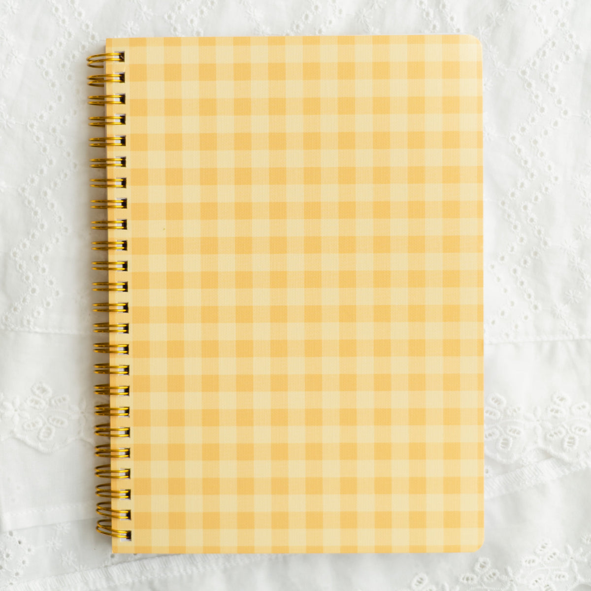 A spiral-bound notebook with a yellow and white gingham checkered cover, placed on a textured white fabric background.