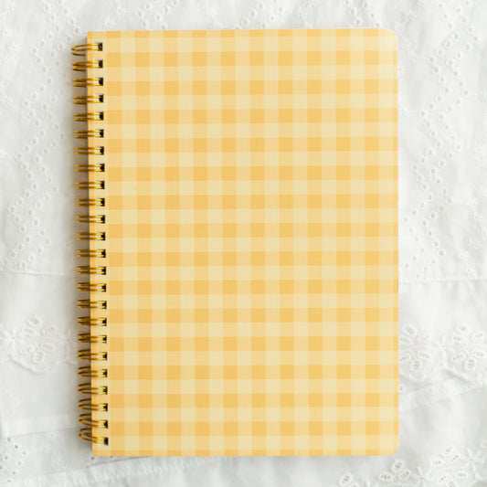 A spiral-bound notebook with a yellow and white gingham checkered cover, placed on a textured white fabric background.