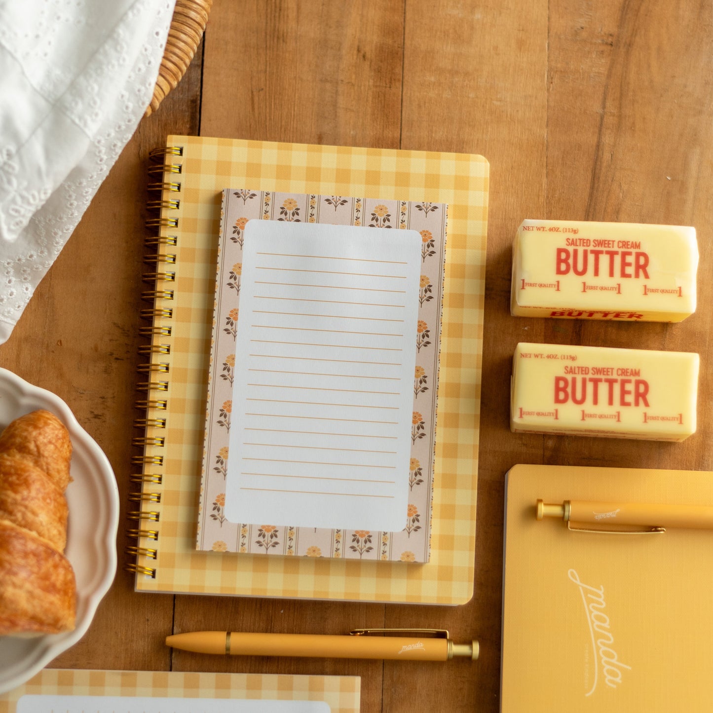 A styled flat-lay of a yellow gingham spiral notebook with a lined paper insert on a wooden tabletop, alongside butter sticks, a pen, another notebook, and a croissant.
