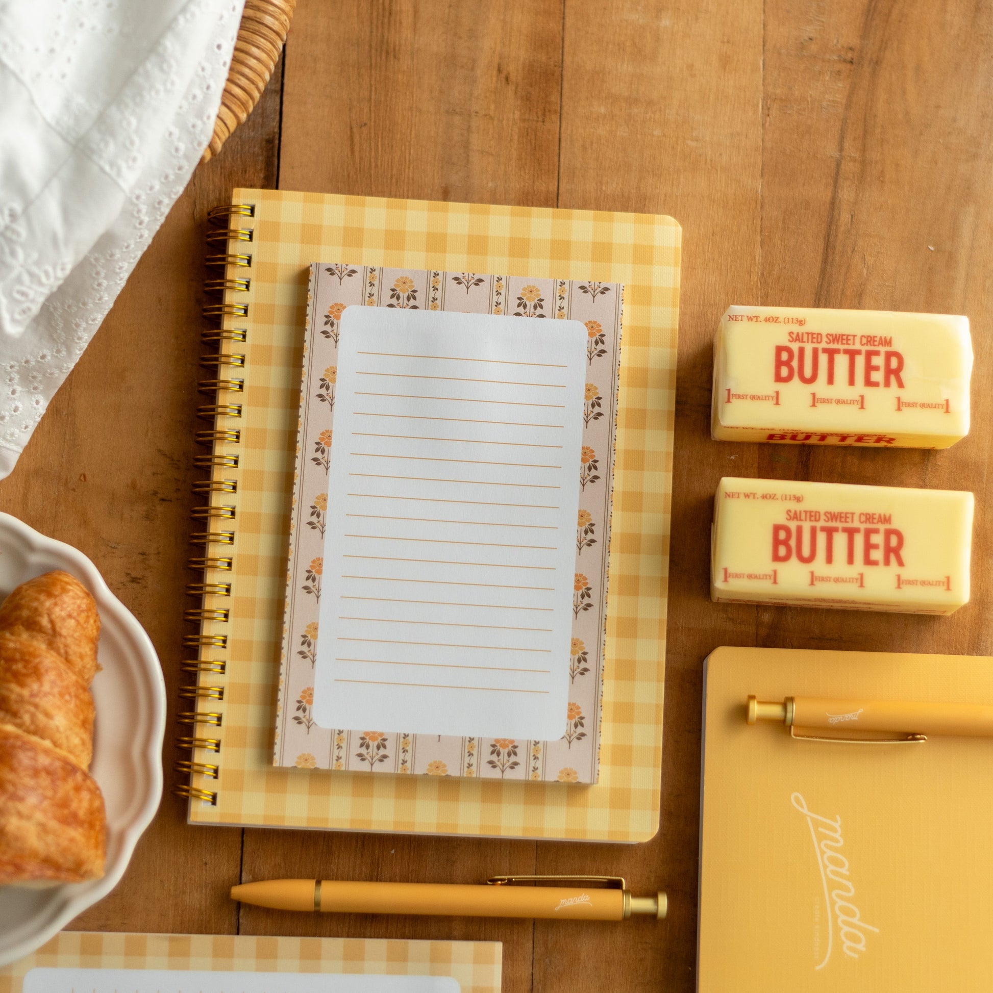 A styled flat-lay of a yellow gingham spiral notebook with a lined paper insert on a wooden tabletop, alongside butter sticks, a pen, another notebook, and a croissant.