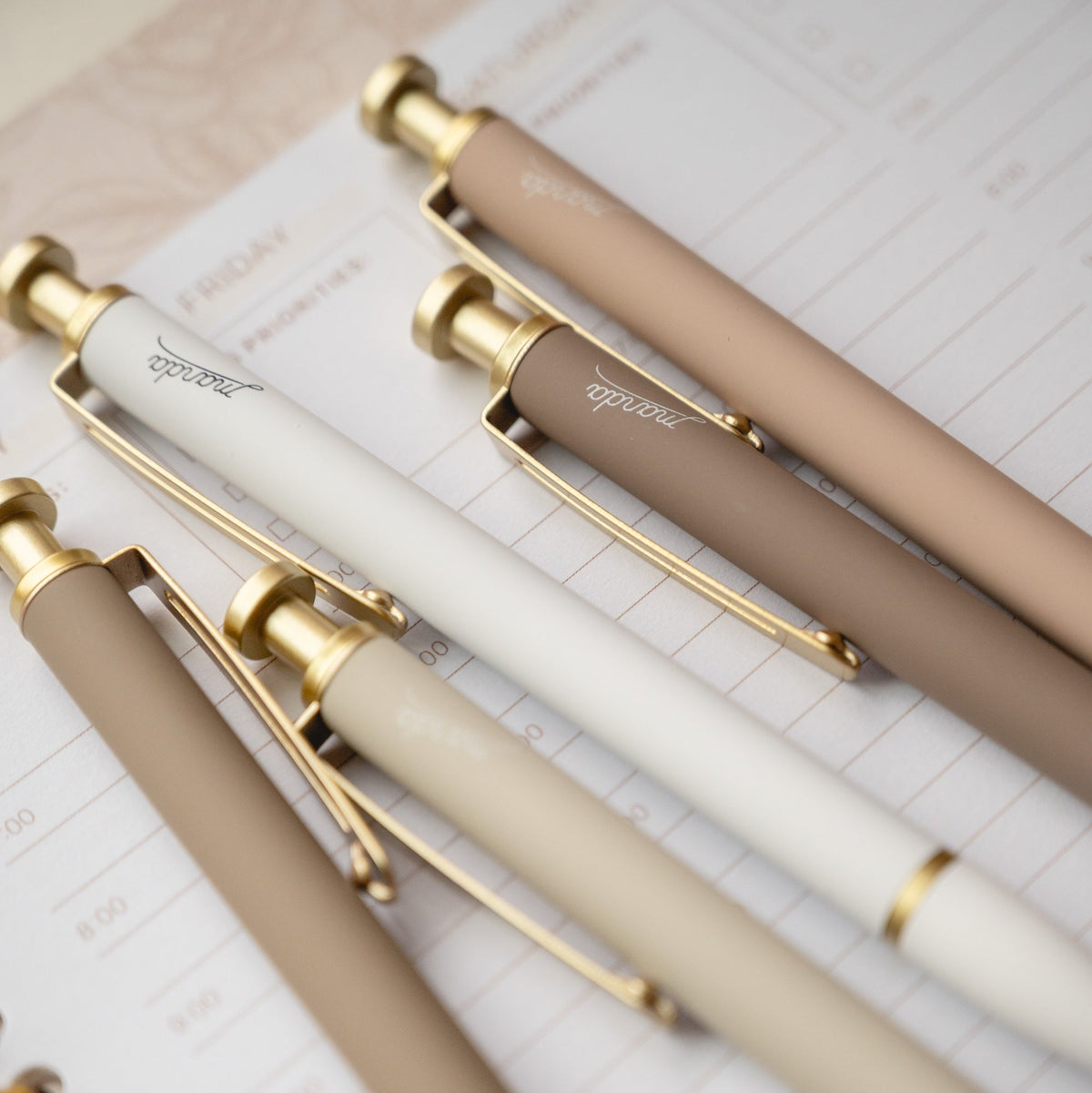 Five elegant ballpoint pens in neutral tones of beige, cream, and taupe, accented with shiny gold metal details, arranged diagonally on an open lined notebook.