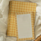 A yellow and white gingham spiral notebook and matching lined notepad resting on white lace fabric inside a wicker basket.