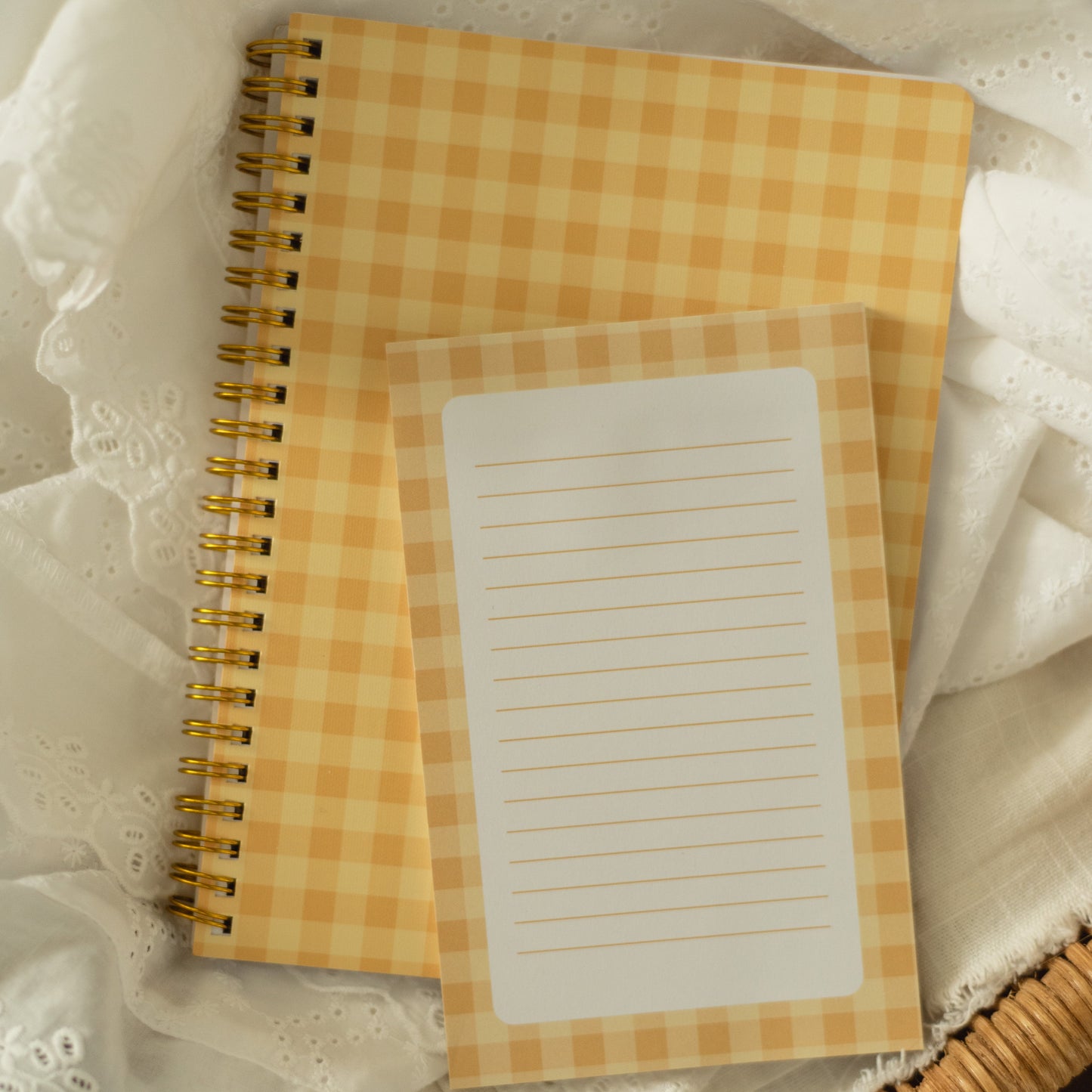 A yellow and white gingham spiral notebook and matching lined notepad resting on white lace fabric inside a wicker basket.