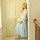 A woman stands barefoot in a doorway wearing a light blue midi dress with a white lace long-sleeve overlay and holding a yellow patterned clutch.