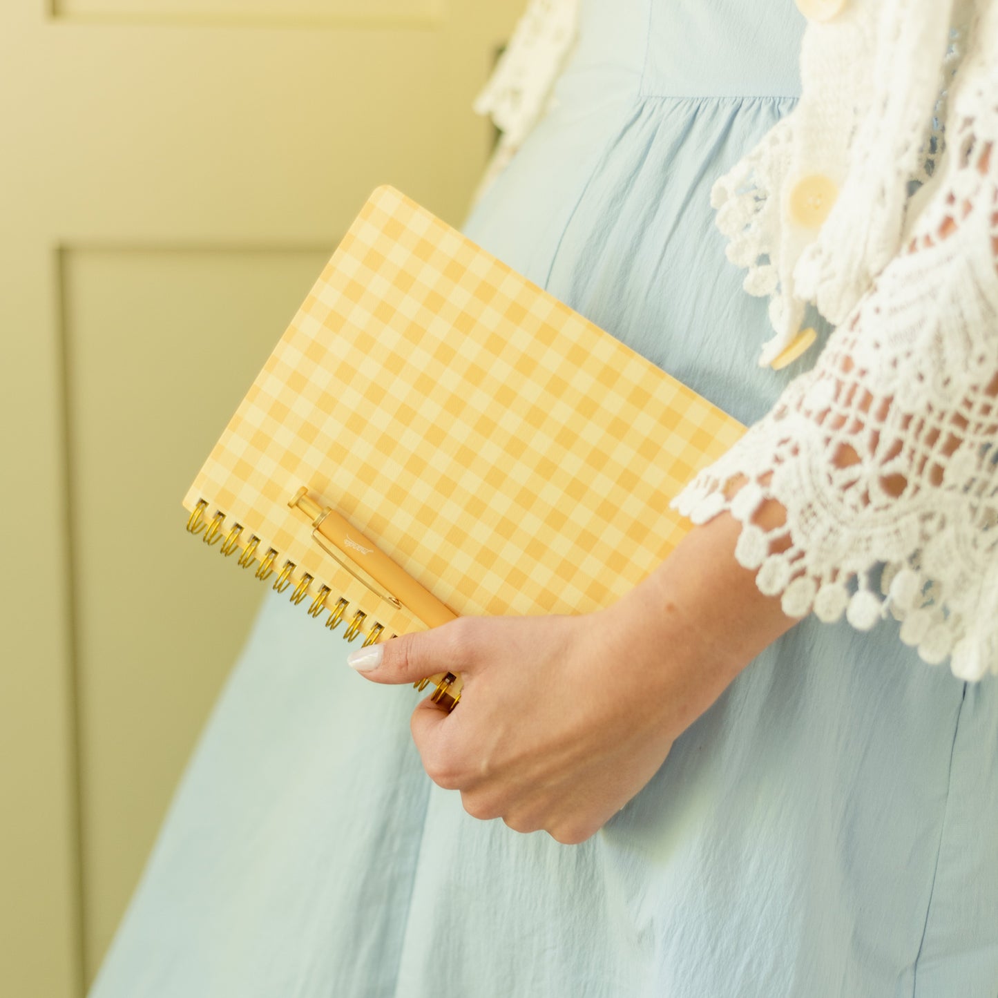 A person in a light blue dress holding a yellow gingham spiral notebook with a gold pen, standing indoors against a pale yellow paneled door.