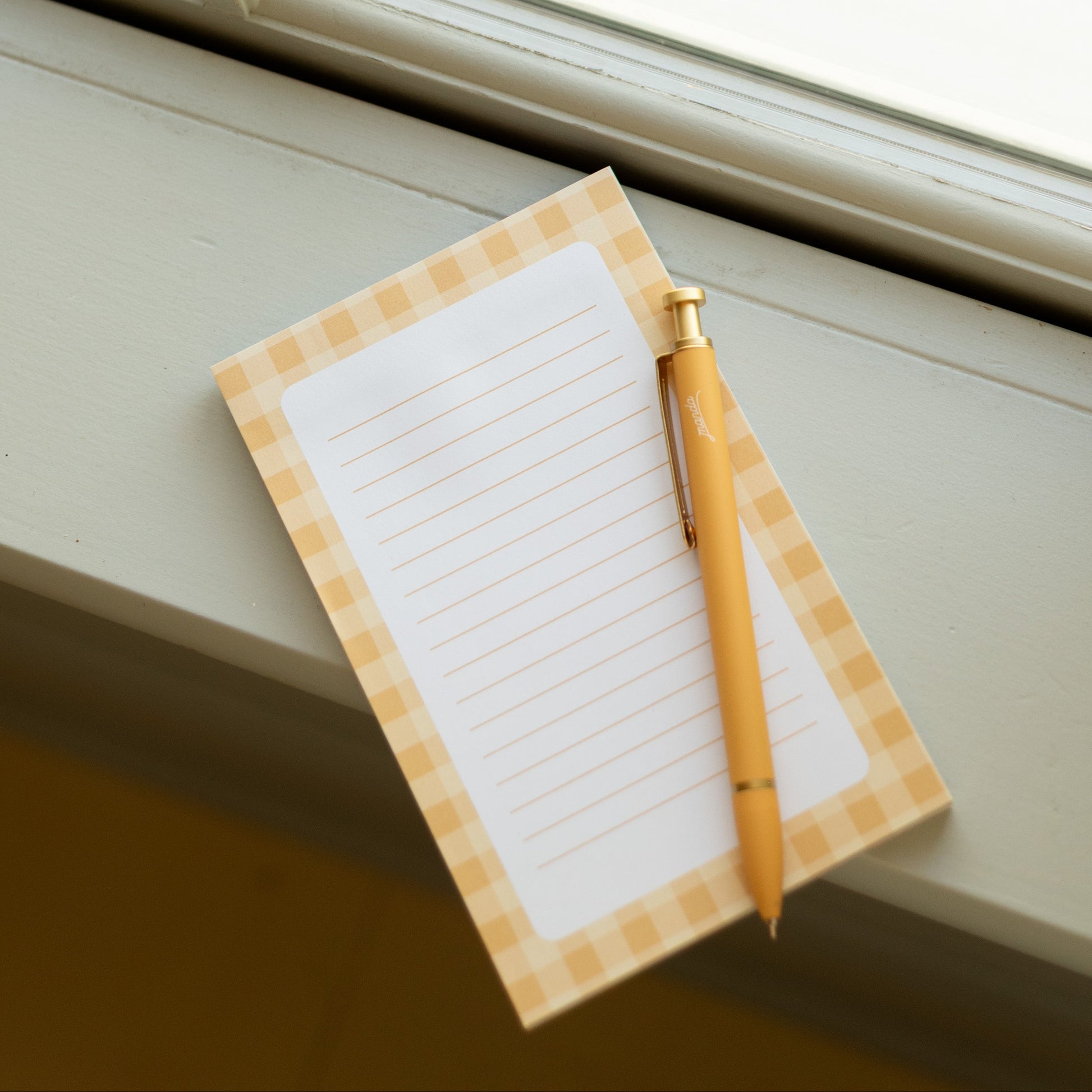 A small lined notepad with a yellow-and-white gingham border rests on a windowsill next to a gold pen. The background shows a softly blurred outdoor view through a white-framed window.