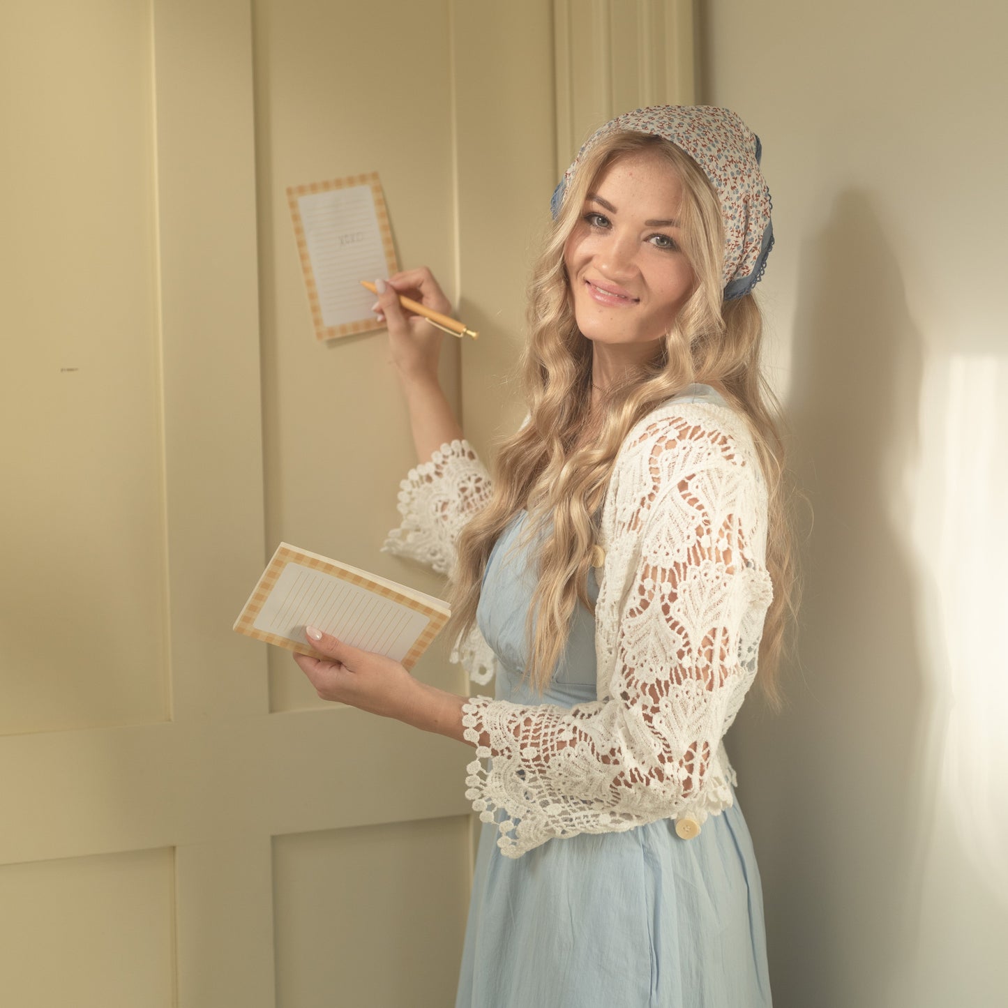 A woman with long blonde hair stands in a room wearing a light blue dress with a white lace bodice and a patterned bonnet. She is holding two small sketchbooks and a pencil, looking to the side with a smile.
