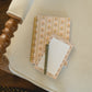 A cozy flat-lay of a floral patterned spiral notebook and matching lined notepad with a green pen, resting on a cream-colored armchair over a dark wood floor.
