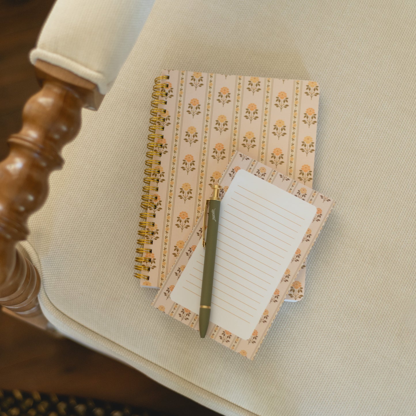 A cozy flat-lay of a floral patterned spiral notebook and matching lined notepad with a green pen, resting on a cream-colored armchair over a dark wood floor.