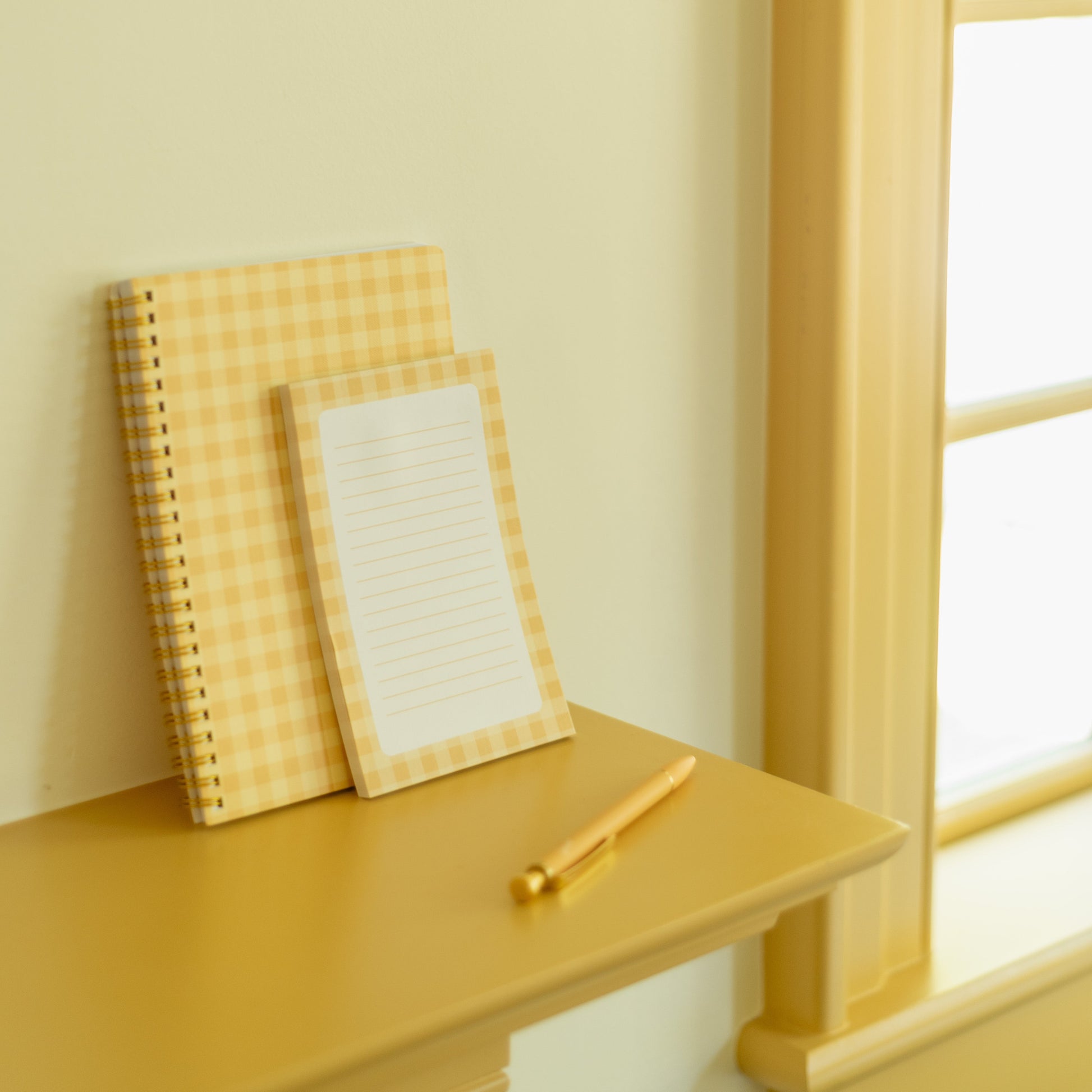 A yellow and white gingham spiral notebook and a matching lined notepad with a gold pen, styled on a yellow wooden mantel next to a window.