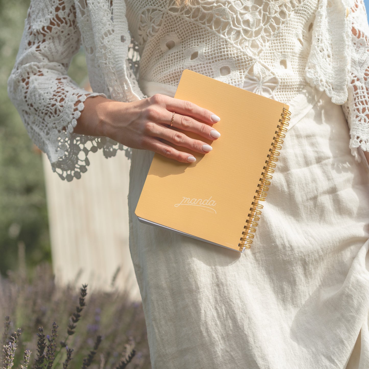 A person in a cream lace top and linen dress holds a mustard yellow spiral notebook with gold rings in a sunlit outdoor setting with blurred lavender in the foreground.