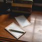A stack of vintage books sits on a wooden windowsill next to a lamp, with two small lined notepads and a gold pen placed in front near an open window.
